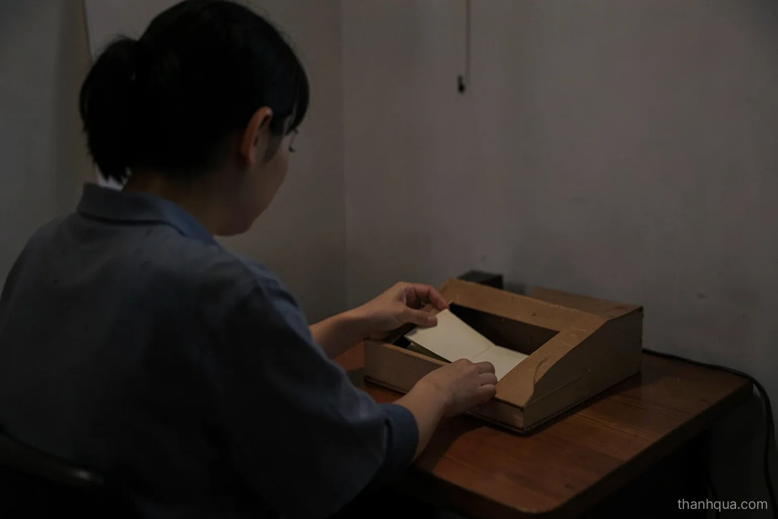 A young Vietnamese office worker seen from behind at a small rented room desk at dusk, dropping a folded envelope into a plain wooden box beside a closed notebook